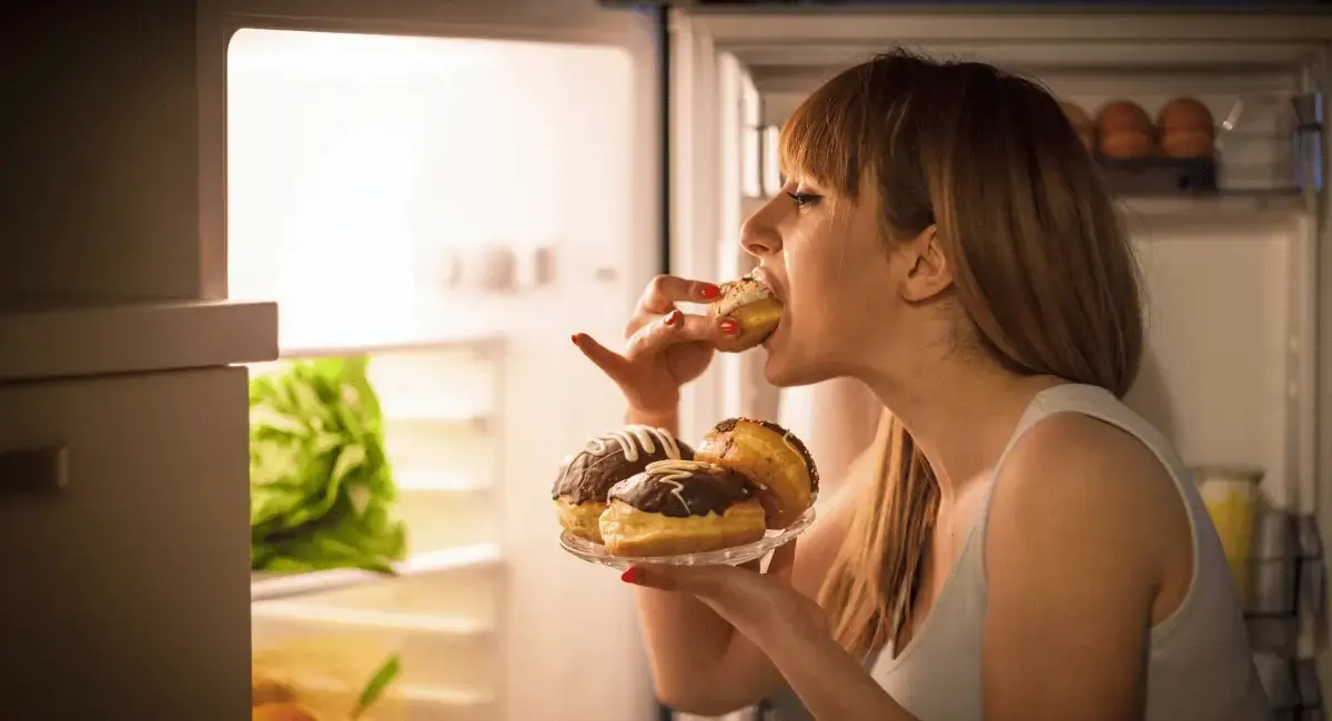 Frau steht gestresst vor dem offenen Kühlschrank und beißt in Donut und hat noch 2 weitere in der Hand.